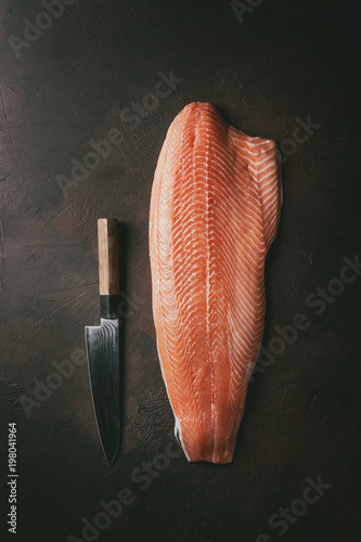 Whole raw uncooked salmon fillet with chef's knife over dark brown texture background. Top view, space.
