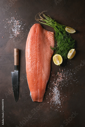 Fotografie Whole raw uncooked salmon fillet server with green dill, lemon, salt, pepper, chef's knife over dark brown texture background