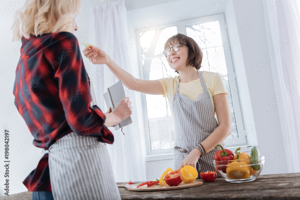 Try it. Joyful nice happy woman standing at the table and smiling while feeding her girlfriend Try it. Joyful nice happy woman standing at the table and smiling while feeding her girlfriend