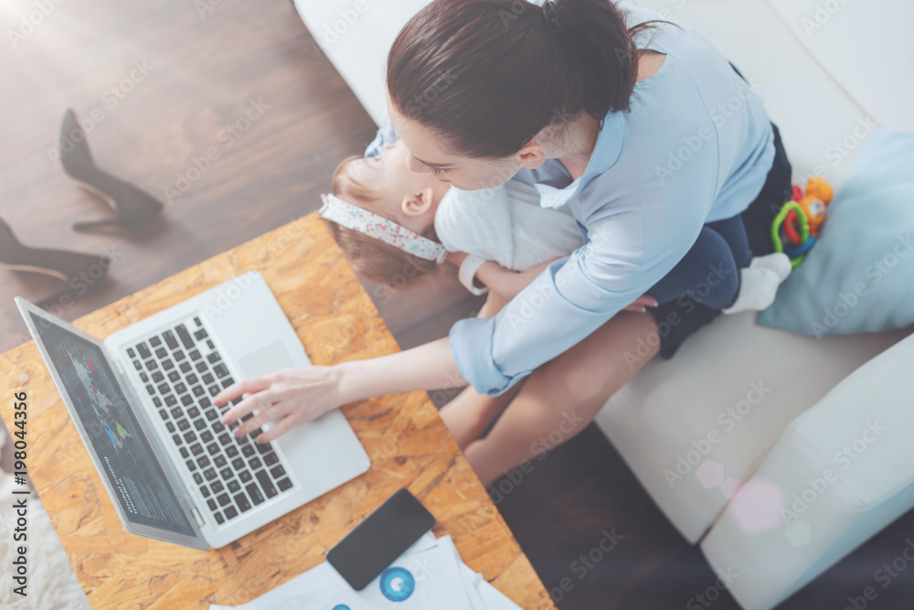 Working mother. Top view of a nice intelligent pleasant woman sitting on the sofa and working while being at home with her child Working mother. Top view of a nice intelligent pleasant woman sitting on the sofa and working while being at home with her child
