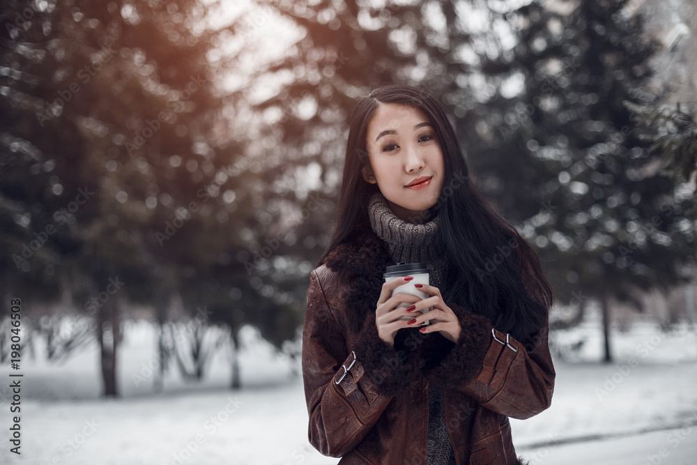 Portrait of young beautiful smiling Asian girl woman in brown knitted sweater and boho leather coat walking outdoors in winter fir park holding a take away cup of coffee or tea Portrait of young beautiful smiling Asian girl woman in brown knitted sweater and boho leather coat walking outdoors in winter fir park holding a take away cup of coffee or tea