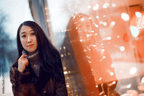 Portrait of young beautiful smiling Asian girl woman in brown knitted sweater and boho leather coat walking outdoors in winter street with magic lights decorations and glass windows