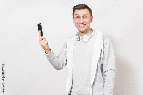 Young handsome smiling man student in t-shirt and light sweatshirt with headphones around neck holds mobile phone in hand and rejoices in studio on white background. Concept of communication, emotions