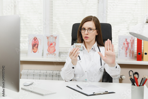 Принти на полотні Female doctor sitting at desk holding bundle of dollars cash money, say No to bribe, showing stop gesture with palm in office in hospital