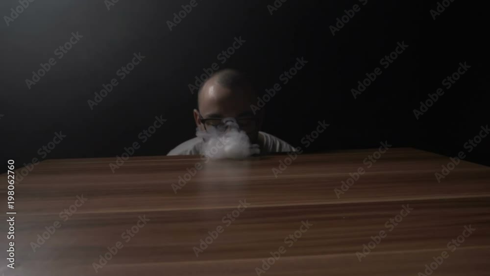 Young hipster teenager boy performing vaping tricks while making a steam wave on the table and then a tornado using his hand in slow motion