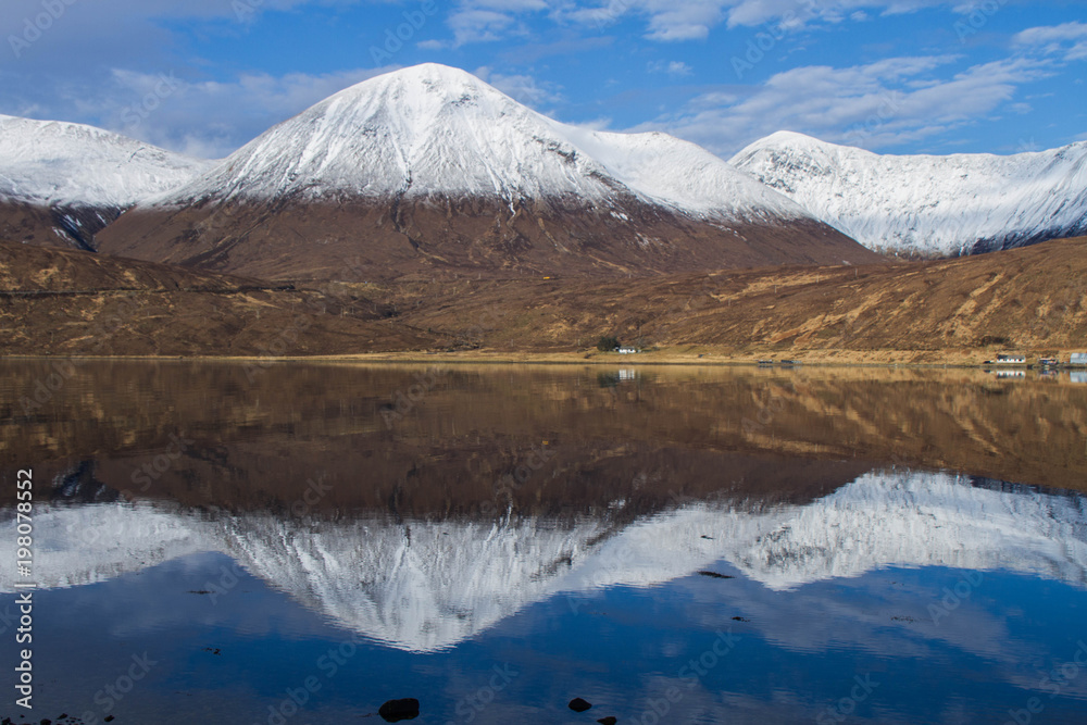 Snow covered peaks of a mountain range and its reflections in Loch Ainort with a little hut standing at the shore of the loch, on the Isle of Skye in the Scottish Highlands