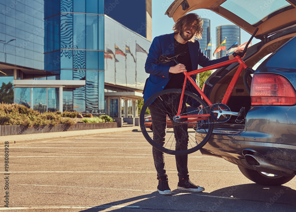 Obraz premium A bearded fashionable bearded male with long hair puts the bicycle in the car's trunk.