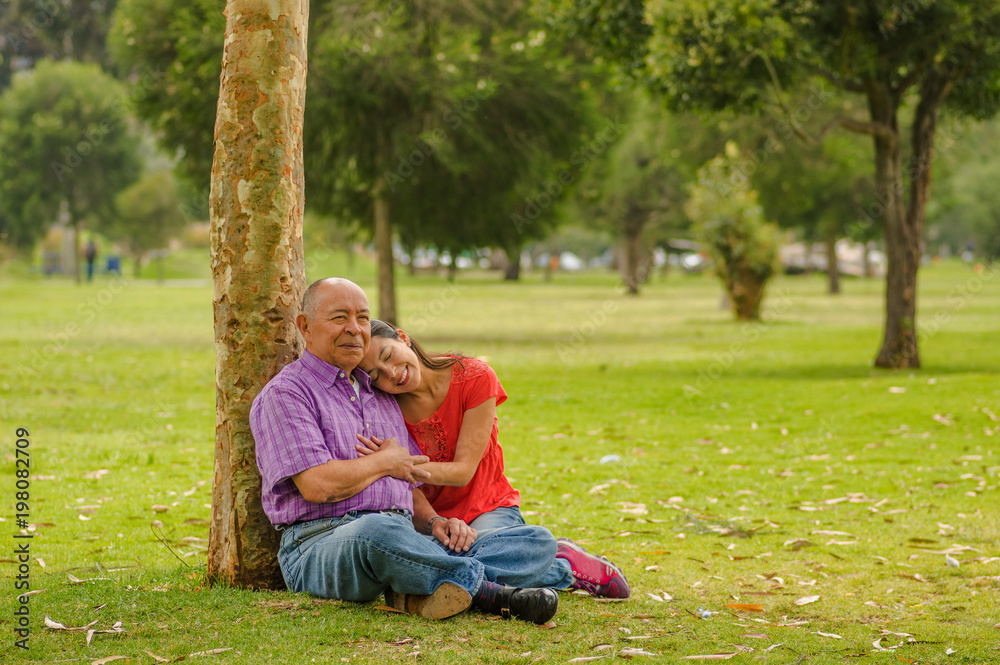 Outdoor view of daughter hugging her father with love at outdoors sitting in the grass, in the park Outdoor view of daughter hugging her father with love at outdoors sitting in the grass, in the park