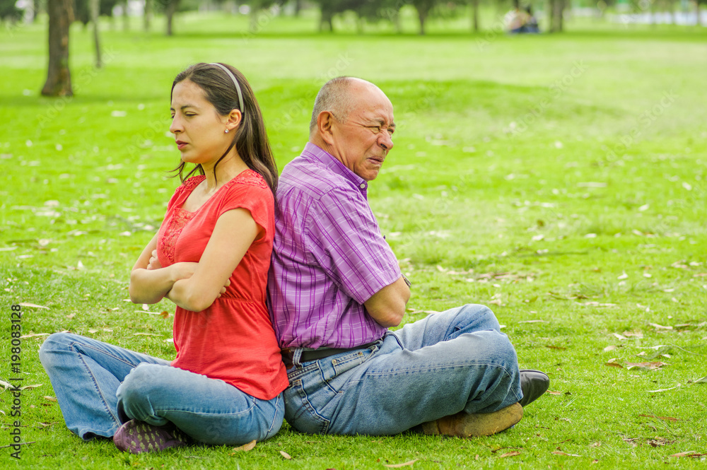 Outdoor view of father and daughter sitting in the grass and back to back each other at outdoors, in the park