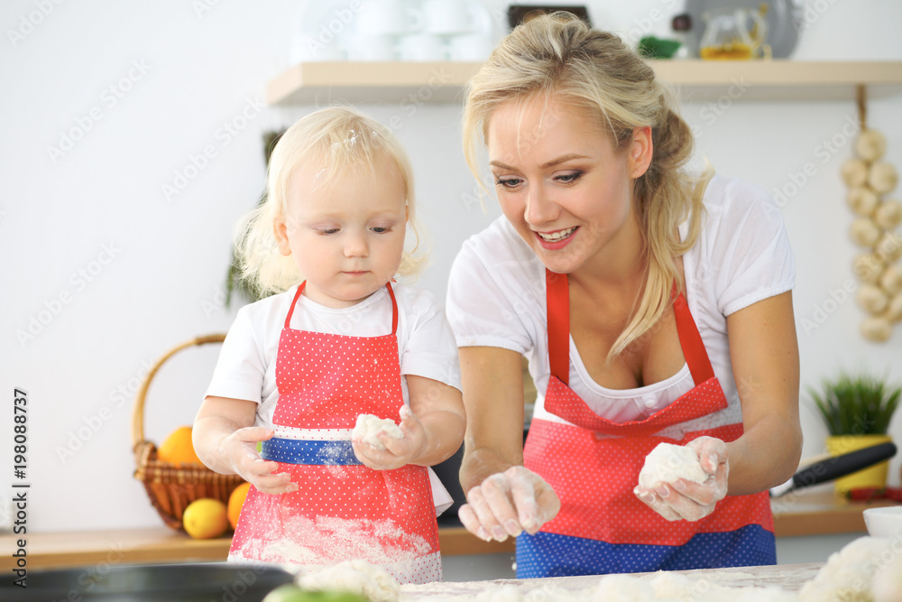 Little girl and her blonde mom in red aprons  playing and laughing while kneading the dough in the kitchen. Homemade pastry for bread, pizza or bake cookies
