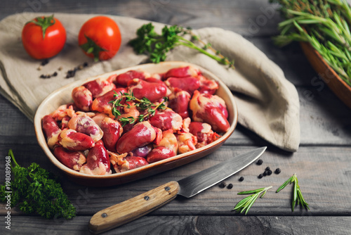 Fresh raw chicken hearts in a ceramic bowl. Seasonings for chicken hearts, thyme, rosemary, parsley, pepper, tomatoes. Rustic wooden gray background.