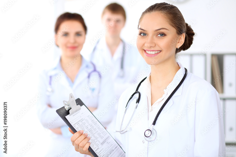 Happy female doctor keeping medical clipboard while medical staff are at the background Happy female doctor keeping medical clipboard while medical staff are at the background