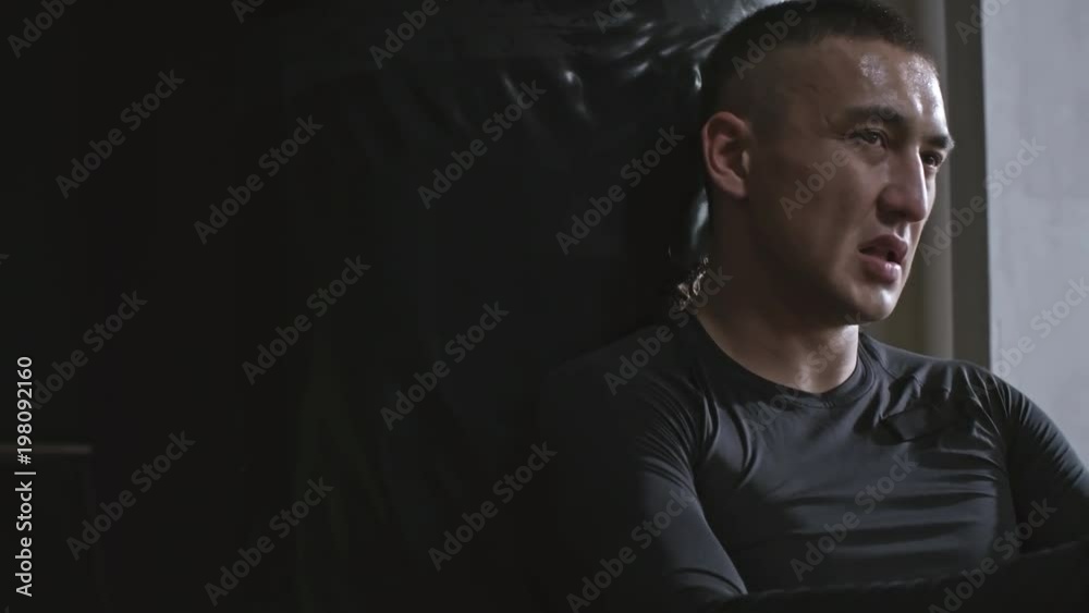 Medium shot of exhausted man relaxing after hard workout at the gym, sitting and leaning on punching bag