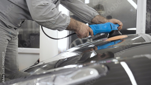 A man in a special suit polishes a gray car body, a tool for polishing cars, into a workshop.