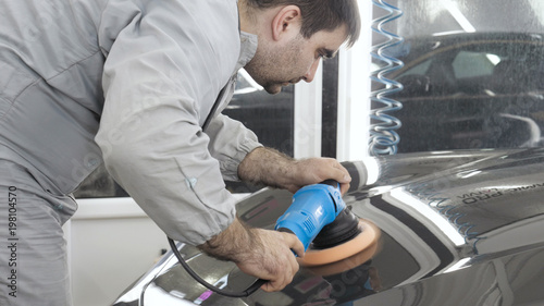 A man in a special suit polishes a gray car body, a tool for polishing cars, into a workshop.