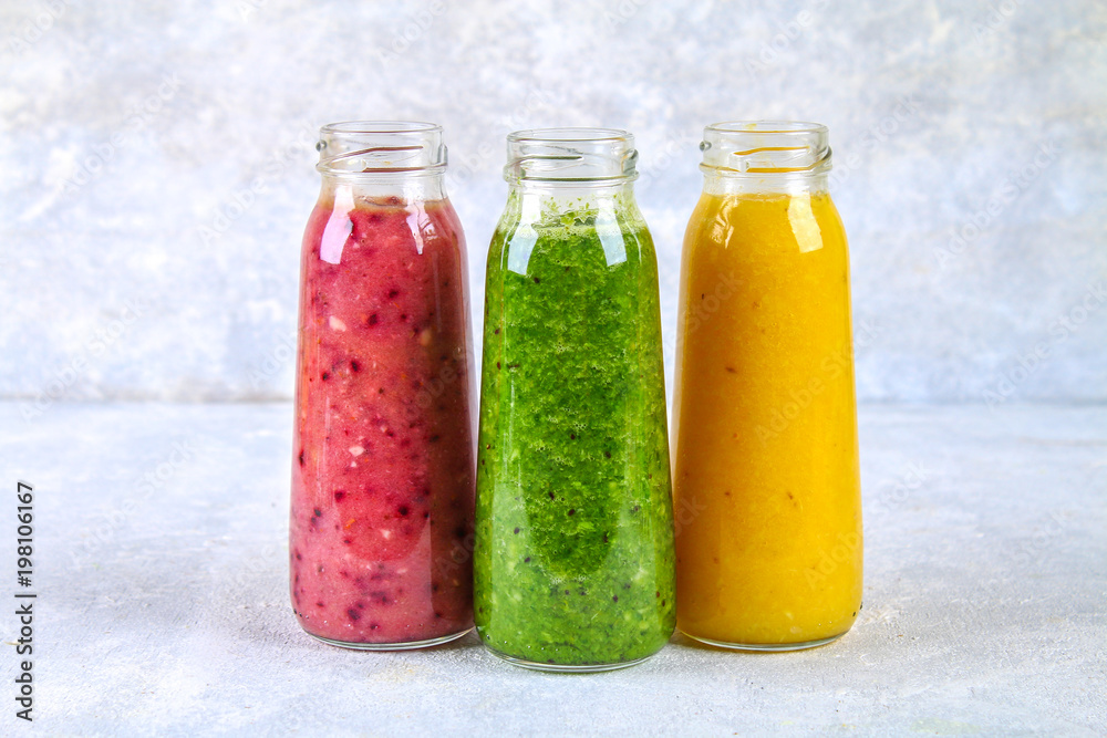 Green, yellow, purple smoothies in bottles of berries, greens, oranges on a white board on a gray table. Green, yellow, purple smoothies in bottles of berries, greens, oranges on a white board on a gray table.