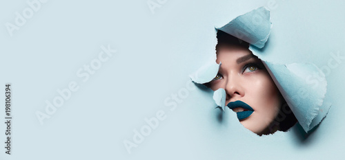 the face of a young beautiful girl with a bright make-up and puffy blue lips peers into a hole in blue paper.