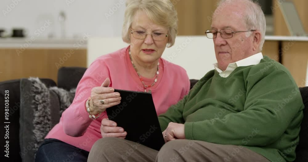 Elderly couple sit on sofa of beautiful contemporary home attempting to use a computer tablet for the first time Elderly couple sit on sofa of beautiful contemporary home attempting to use a computer tablet for the first time