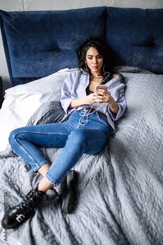 A beautiful brunette listens to music in headphones in a room near the sofa, is dressed in a shirt and blue jeans, blue