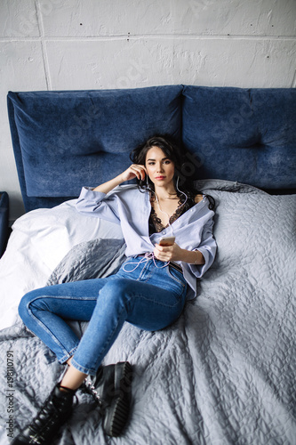 A beautiful brunette listens to music in headphones in a room near the sofa, is dressed in a shirt and blue jeans, blue