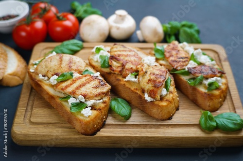Tasty crunchy italian snack bruschetta with grilled chicken, garnished with ricotta and basil on wooden board