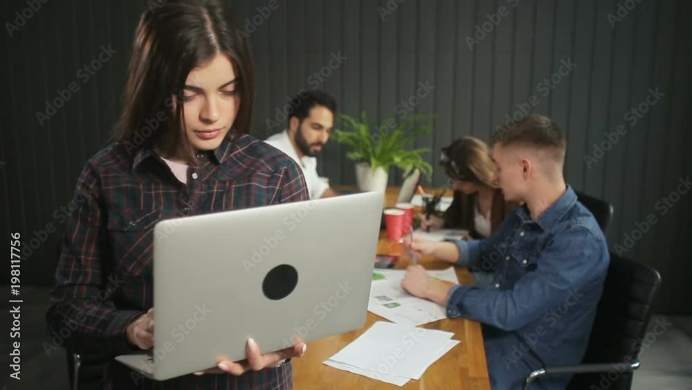 Lovely brunette typing on silver laptop as standing before busy team of specialists, young team of professionals using gadgets while carrying out the task, indoor shot in dark grey office Lovely brunette typing on silver laptop as standing before busy team of specialists, young team of professionals using gadgets while carrying out the task, indoor shot in dark grey office