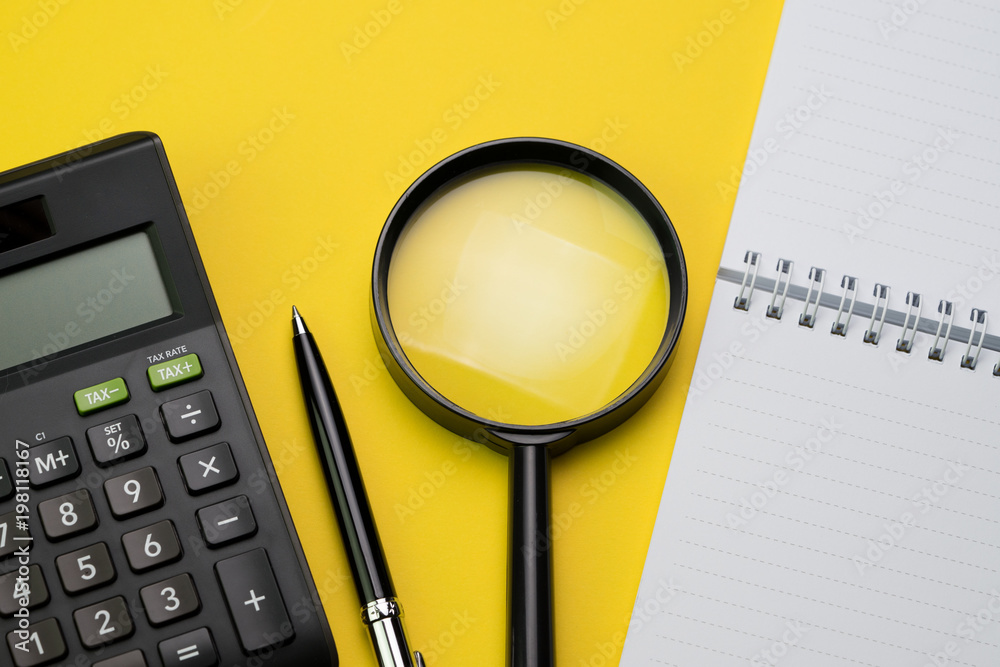 Flat lay or top view of black pen, calculator, magnifying glass and white  notepad on vivid yellow background table with blank copy space, math, cost,  tax or investment calculation Stock Photo