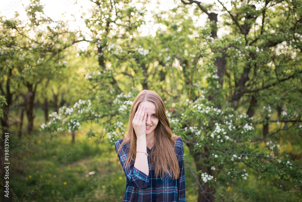 A young beautiful girl with long hair in a blue shirt in a cage into the blossoming apple tree. The girl closed one eye with her hand. Spring A young beautiful girl with long hair in a blue shirt in a cage into the blossoming apple tree. The girl closed one eye with her hand. Spring