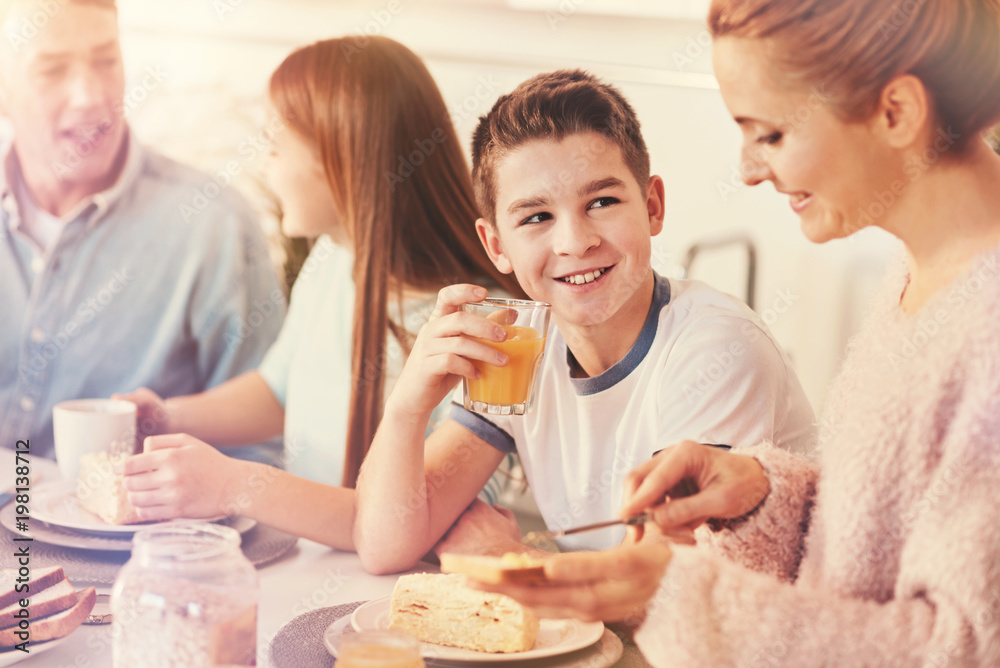 Good mood. Positive teenager keeping smile on his face and putting elbow on the table while holding glass with orange juice Good mood. Positive teenager keeping smile on his face and putting elbow on the table while holding glass with orange juice