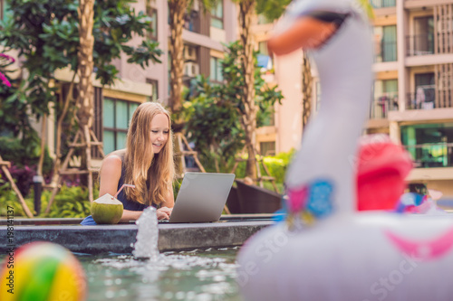 Young female freelancer sitting near the pool with her laptop in the hotel browsing in her smartphone. Busy at holidays. Distant work concept. Copy space for your text photo