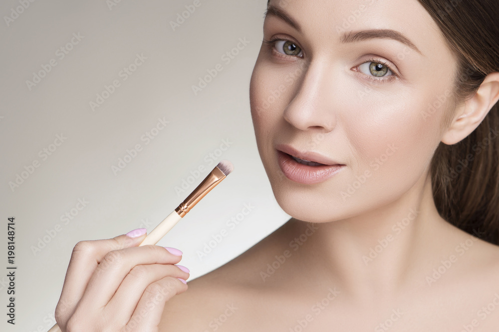 Obraz premium Beautiful brunette caucasian young woman prepare herself, applying lipstick on her lips with a brush. Clean, fresh, natural, flawless skin. Soft smile on her face. Close up on a neutral background