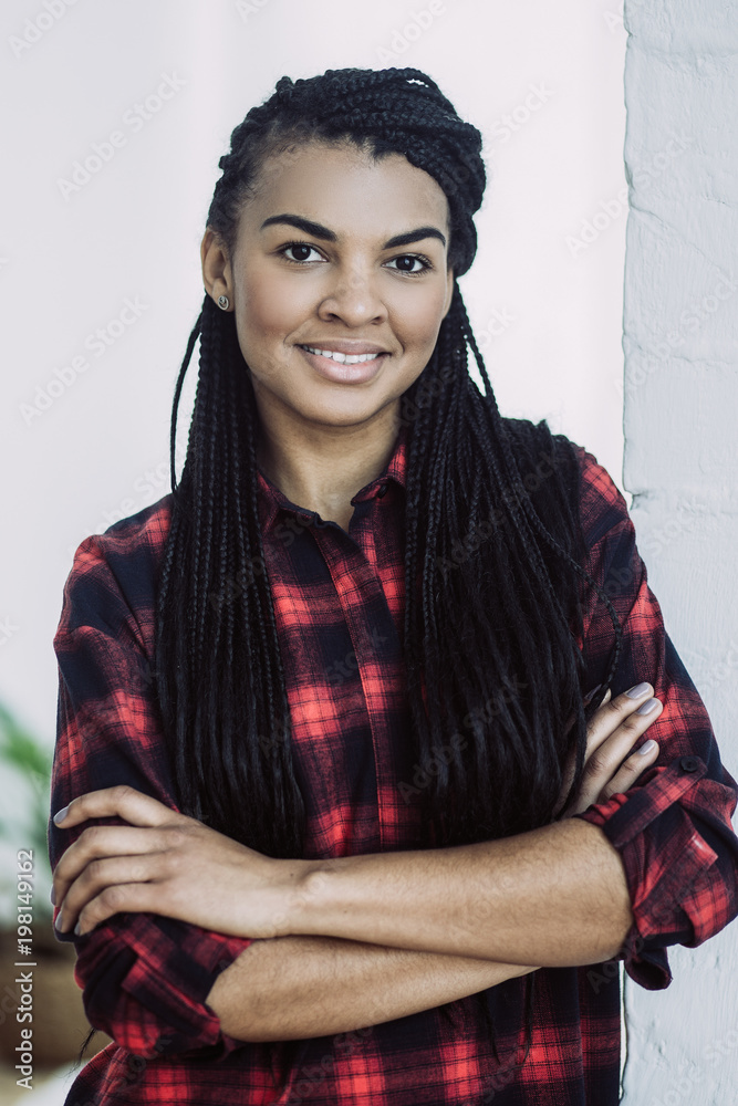 Portrait of happy stylish young African-American woman wearing red checked shirt standing with folded arms, looking at camera and smiling at home Portrait of happy stylish young African-American woman wearing red checked shirt standing with folded arms, looking at camera and smiling at home