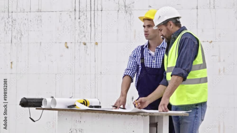 Two male engineers standing at table and using building blueprint to discuss project details when working at construction site Two male engineers standing at table and using building blueprint to discuss project details when working at construction site