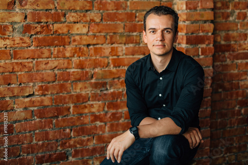 Cute young man in black shirt sitting on chair in studio near brick wall and looking at camera.