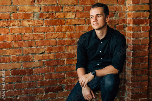 Cute young thoughtful man in black shirt sitting on chair in studio near brick wall.