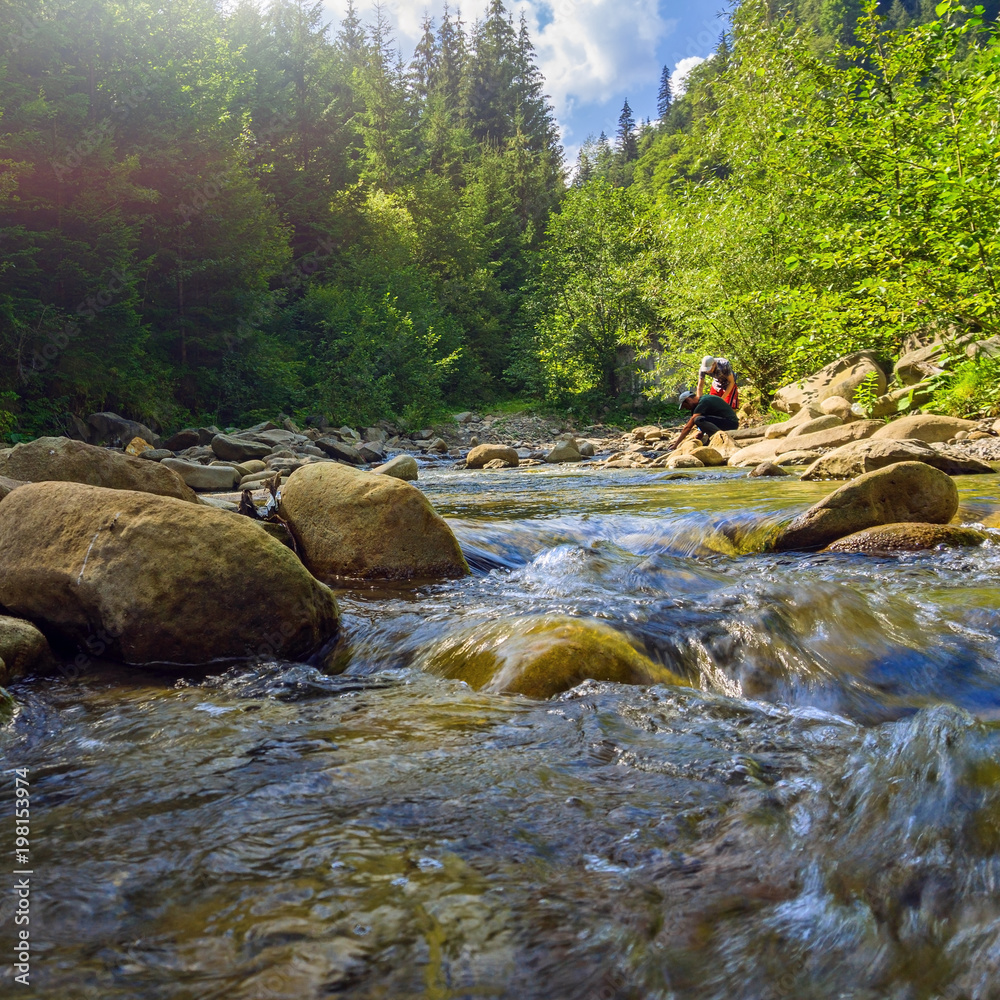 Two tourists are fetching water from mountain river in summer Carpathians. Landscape of forest stream in mountains with people.