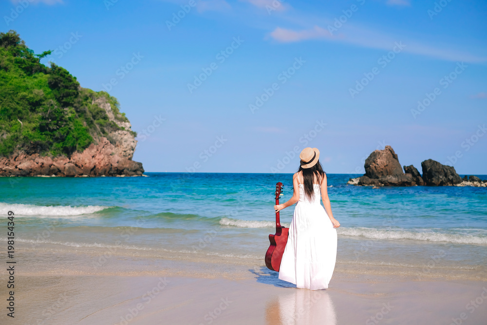 woman traveler in white dress standing and holding a guitar on the beach during her holiday.