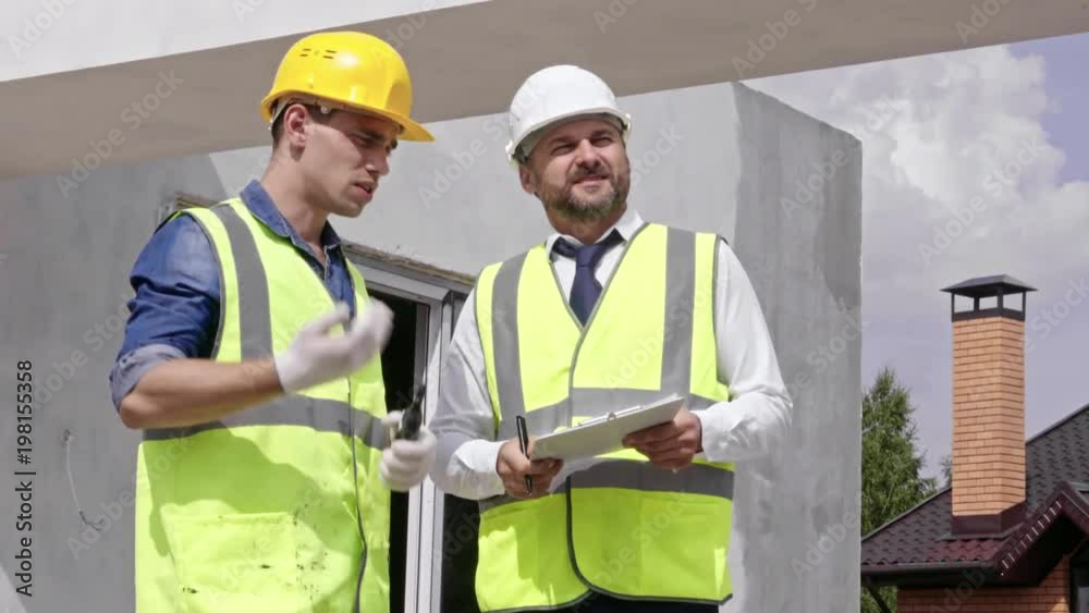 Medium shot of two engineers wearing hard hats and safety vests, one holding clipboard, looking at building under construction and talking