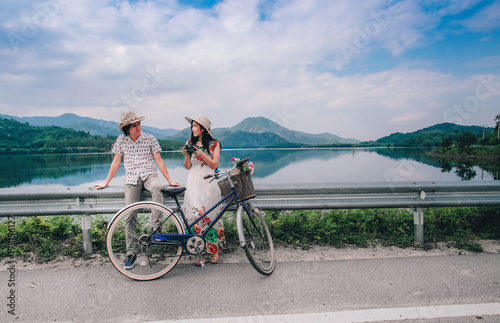 couple traveler standing beside bicycle and happy for view of nature near the lake on vacation background is mountain.