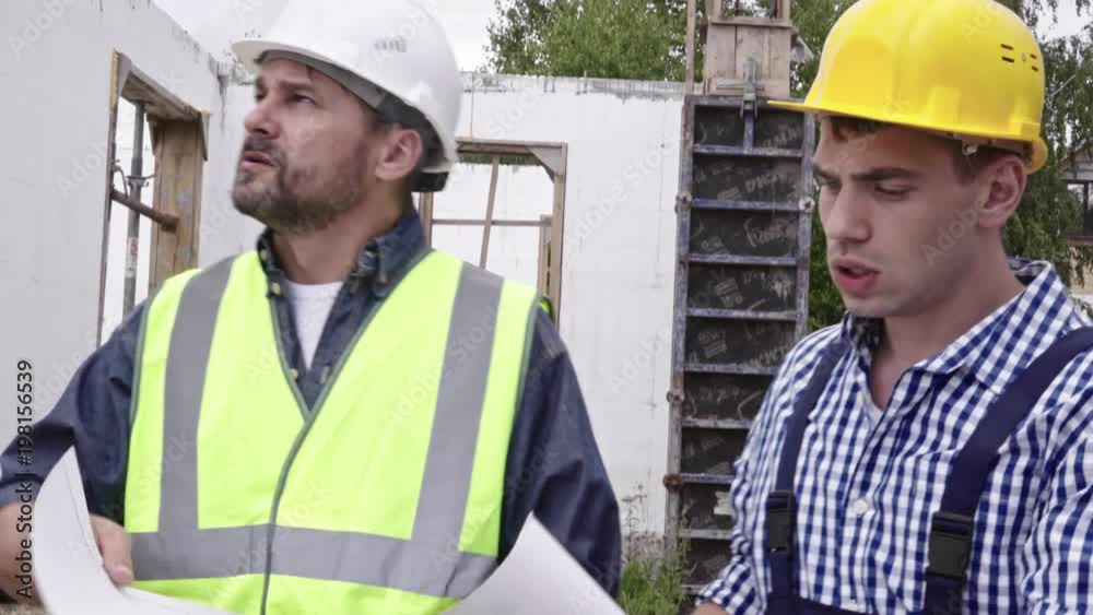 Two male engineers in hard hats discussing blueprint of future building and talking about work process at construction site Two male engineers in hard hats discussing blueprint of future building and talking about work process at construction site