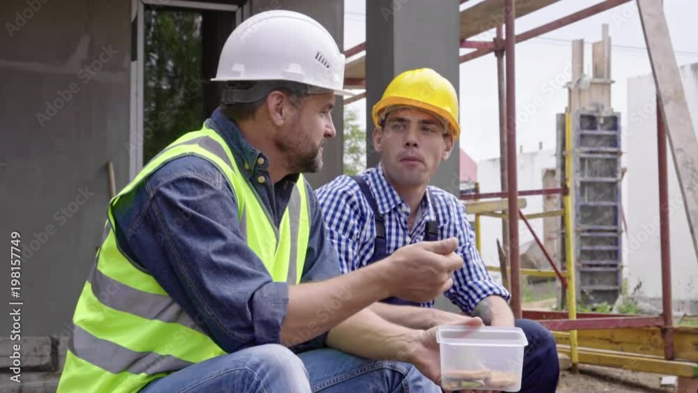 Medium shot of two male engineers in hard hats taking break from work, talking to each other and eating food from lunch box Medium shot of two male engineers in hard hats taking break from work, talking to each other and eating food from lunch box