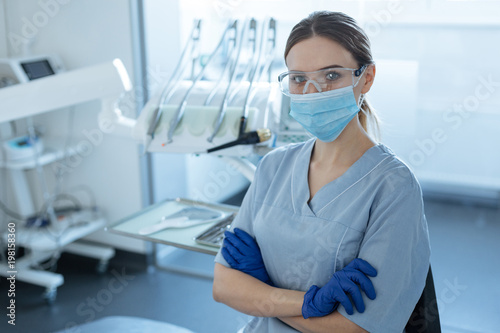 Ready for treatment. Pretty young female dentist wearing safety goggles, face mask and rubber gloves while posing for the camera and folding her hands across her chest
