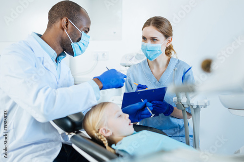 Important data. Upbeat young male dentist dictating his observations to his pleasant nurse while examining the teeth of his little patient