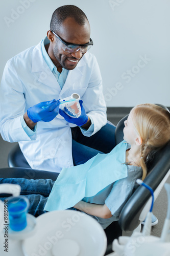 Correct technology. Charming young male dentist smiling at his little patient while showing her how to brush teeth and remove dental plaque correctly