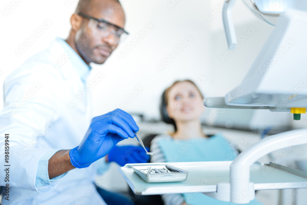 Fototapeta premium Of best quality. The focus being on the hand of a pleasant young male dentist putting a mouth mirror on a tray, having used it during the examination of his female patient