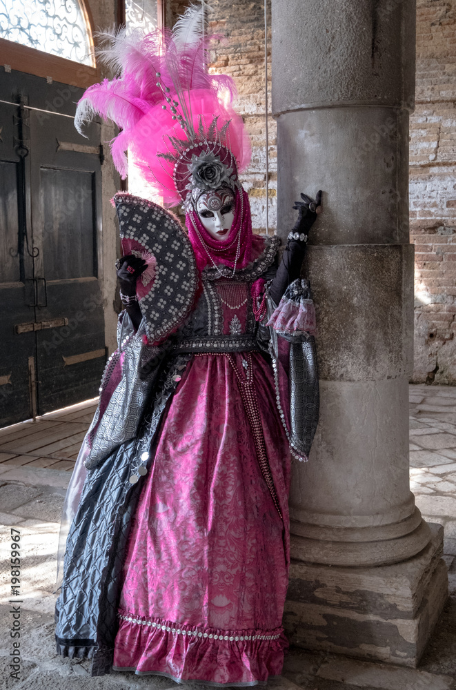  Masked lady in ornate pink costume dress and mask standing next to pillar at Venice Carnival. Lady has feathered plumes, holds a decorated fan and is wearing black gloves