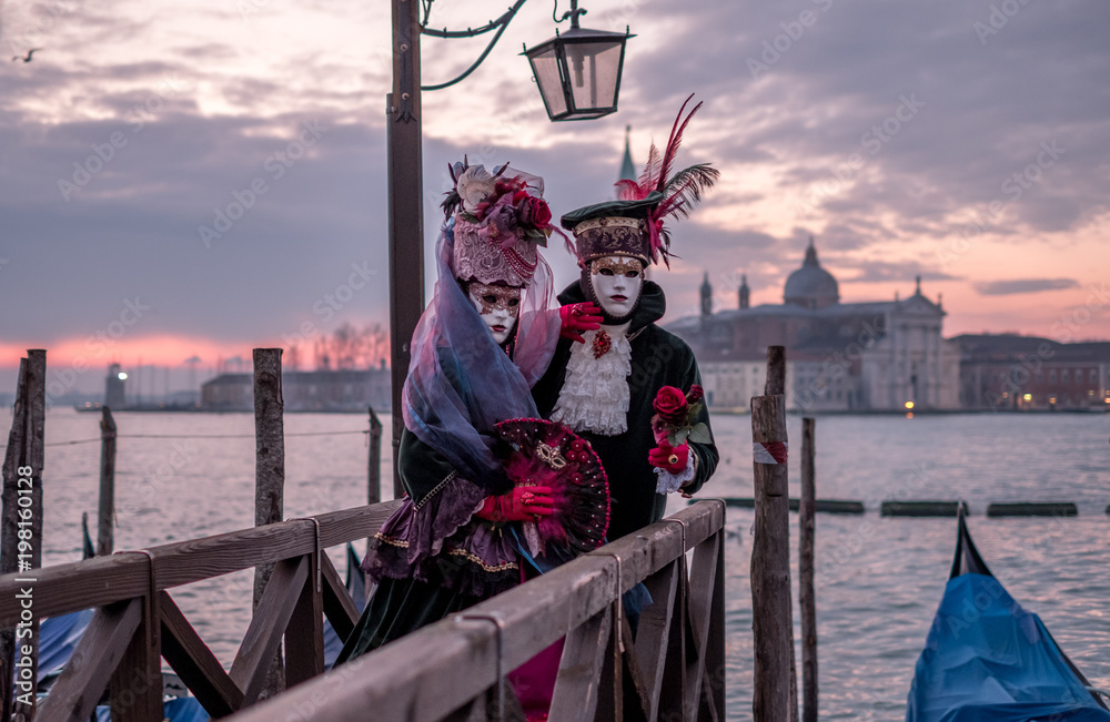 Φωτογραφία Romantic couple in traditional costume and masks standing with back to the Grand Canal, gondolas and San Giorgio in the background, during the Venice Carnival