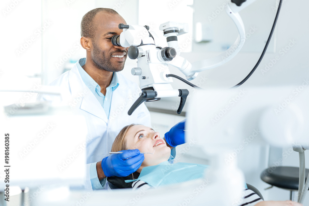 Examining thoroughly. Upbeat male dentist using a professional microscope and checking for cracks in the teeth of his patient Examining thoroughly. Upbeat male dentist using a professional microscope and checking for cracks in the teeth of his patient