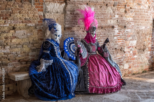 Two women in masks and ornate blue and pink costumes and decorative fans, sitting on a stone bench against an old brick wall. 