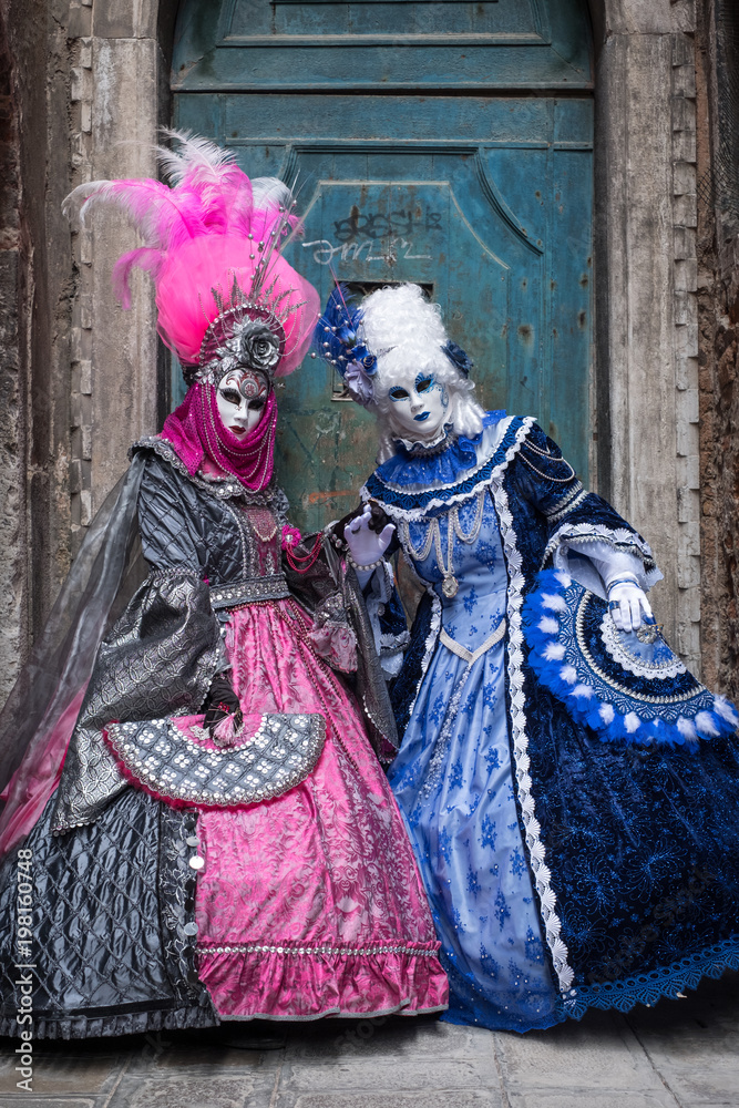 Two women in masks and ornate blue and pink costumes standing in front of an old blue door at the end of an alley in Venice during the carnival (Carnivale di Venezia).